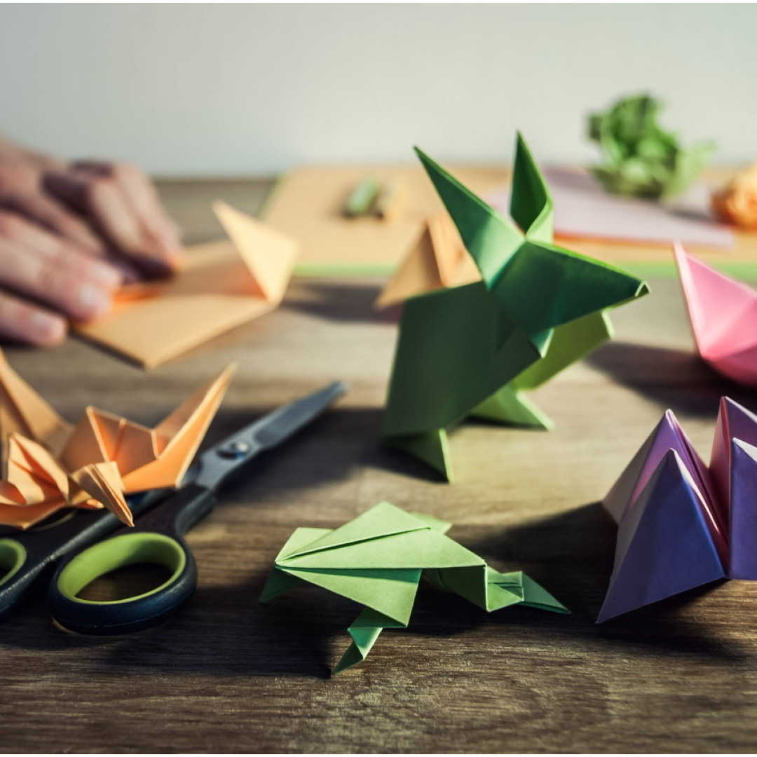 Origami figures, scissors and pencils on wooden table, in the background hands folding colored paper